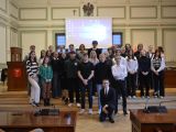 W sali posrodku na podwyższeniu stoi duża grupa uczniów liceum. Za nimi na ścianie wisi ekran projekcyjny.