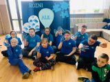 Uczniowie w niebieskich koszulkach siedzą na podłodze z uniesionymi dłońmi pomalowanymi na niebiesko