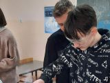 Chłopak w oklarach robi doświadczenie chemiczne na stole. Obok niego stoi chłopak, który się temu przygląda.