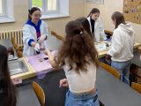 W sali pośrodku przy długim roboczym stole stoją cztery dziewczyny z liceum, wykonują doświadczenia chemiczne. Dwie...