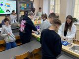 W sali pośrodku przy długim roboczym stole stoją dwie dziewczyny z liceum w białych fartuchach, wykonują doświadczenia...