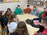 Przy stoliku siedzi dziesięcioro uczniów szkoły podstawowej i przygląda się jak dziewczyna wykonuje doświadczenie chemiczne.