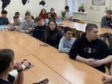 W sali lekcyjnej przy długim stole siedzą uczniowie liceum. Słuchają tego co mówi do nich kobieta.