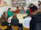 Przy stoliku siedzi dziesięcioro uczniów szkoły podstawowej i przygląda się jak dziewczyna wykonuje doświadczenie chemiczne.