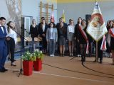 Duża grupa uczniów liceum stoi w trzech rzędach w sali. Z prawej strony uczeń trzyma sztandar, z lewej strony dziewczyna i...