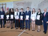 Ośmioro uczniów liceum stoi w rzędzie, w rękach trzymają świadectwa. Obok nich stoi mężczyzna w garniturze.