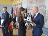 Mężczyzna w garniturze wręcza świadectwo licealistce w okularach. W tle stoi licealista w garniturze, którzy trzyma...