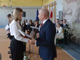 Mężczyzna w garniturze wręcza świadectwo licealistce w białej bluzce. W tle siedzi grupa uczniów liceum.