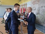 Mężczyzna w garniturze wręcza świadectwo licealiście w garniturze w kratkę. W tle stoi troje uczniów liceum.