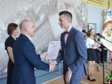 Mężczyzna w garniturze wręcza świadectwo licealiście w garniturze. W tle stoi kobieta i dwoje uczniów liceum.