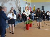 Dziewczyna w białej bluzce podaje ksiązkę mężyczźnie, obok stoi kobieta w okularach. W stronę mężczyzny idzie licalistka w...