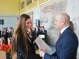 Mężczyzna w garniturze wręcza świadectwo dziewczynie z długimi włosami. W tle stoi kobieta w białej marynarce, a na...