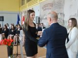 Mężczyzna w garniturze wręcza świadectwo dziewczynie z długimi włosami. Obok stoi kobieta w białej marynarce. W tle na...