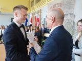 Mężczyzna w garniturze wręcza świadectwo chłopakowi z włosami blond. Obok stoi kobieta w białej marynarce. W tle na...