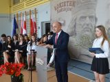 Przy mównicy do mikrofonu przemawia mężczyzna w garniturze, obok stoi licealistka w białej bluzce. W tle na krzesłach...