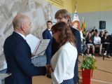 Mężczyzna w garniturze wręcza dyplom kobiecie i mężczyźnie. W tle uczniowie liceum siedzą na krzesłach.