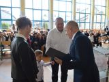 Mężczyna w garniturze wręcza dyplom kobiecie, obk której stoi dziecko i drugi mężczyzna. W tle na krzesłach siedzą ludzie.