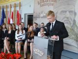 Przy mównicy do mikrofonu przemawia uczeń liceum w garniturze. W tle na krzesłach siedzi grupa uczniów liceum.