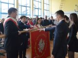 Uczeń liceum trzyma opuszczony sztandar. Wokół sztandaru stoi czworo licealistów z podniesioną do góry ręką i składa...