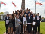 Duża grupa uczniów liceum stoi przy pomniku. W tle flagi biało-czerwone i drzewa.