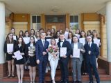 Duża grupa uczniów liceum stoi przed budynkiem wszkoły. Wśród uczniów stoi kobieta z kwiatami.