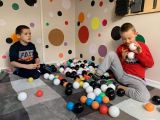 Dwoje uczniów siedząc na szarym kocu bawi się kolorowymi piłkami