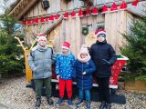 Czworo uczniów szkoły podstawowej w czapkach św. Mikołaja. Za nimi drewniana szopa i czerwone sanie