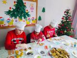 Troje uczniów szkoły podstawowej w czapkach kucharskich ozdabia pierniczki leżące na stole. W tle choinka i...