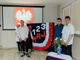 Gimnazjaliści ubrani na galowo na tle tablicy z napisem 1,2,3 maja. W tle wyświetlane projektorem godło Polski