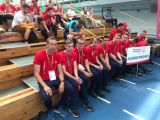 Grupa gimnazjalistów z nauczycielem w czerwonych strojach sportowych siedzi na drewnianej ławce w hali sportowej