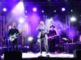 Na scenie stoi mężczyzna w siwym garniturze i śpiewa do mikrofonu. Z tyłu dwaj mężczyźni grają na gitarach. Jest wieczór.