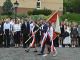 Przed szkołą idzie licealista niosący sztandar szkoły. Obok niego idą dwie licealistki w białych bluzkach. W tle duża grupa...