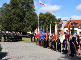 Na rynku miasta w rzędzie stoi siedmiu chłopców trzymających przed soboą sztandary. Z lewej strony stoi grupa młodzieży w...