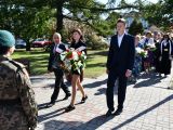 Dziewczyna w czarnej garsonce niesie biało-czerwone kwiaty. Obok niej z lewej strony idzie mężczyzna w garniturze, z prawej...