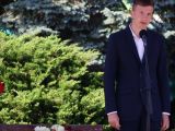 Chłopak w garniturze przemawia do mikrofonu. W tle zielone krzewy.