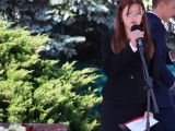 Dziewczyna w czarnej garsonce i białej bluzce śpiewa do mikrofonu. W tle zielone krzewy.