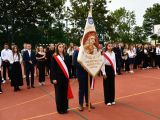 Pośrodku boiska chłopak w garniturze trzyma sztandar. Obok niego stoją dwie uczennice liceum. W tle w rzędach stoją...