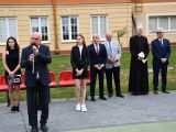 Mężczyzna w garniturze przemawia do mikrofonu. W tle stoją dwie dziewczyny z liceum, mężczyzna w sutannie, trzech mężczyzn...