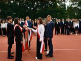 Uczeń liceum trzyma opuszczony sztandar, który podtrzymują dwie dziewczyny. Po prawej stronie sztandaru stoją dwie...