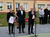 Przy dwóch mikrofonach stoi ddwóch licealistów i mężczyzna, wszyscy są w garniturach. W tle z prawej strony stoją dwaj...