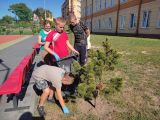 Czworo uczniów na dziedzińcu szkoły. Pochylony chłopiec zbiera papierki leżące na trawniku. Za nim uczeń trzymajacy foliowy...