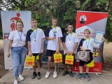 Czterech uczniów w strojach sportowych z medalami na piersi i żóltymi pudełkami w dłoniach. Chłopiec w środku trzyma puchar