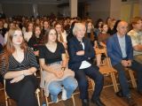 W sali na krzesłach, w rzędach, siedzi duża grupa uczniów liceum. W pierwszym rzędzie między uczniami siedzi starszy...
