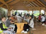 Uczniowie liceum siedzą pod drewnianym zadaszeniem przy dużych drewnianych stołach i jedzą posiłek.