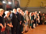 W sali gimnastycznej, w rzędach stoją ludzie w różnym wieku. W tle duże zdjęcia zachodów słońca.