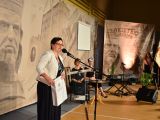 Do mikrofonu przemawia kobieta w białej marynarsce. W ręku trzymam papierową torbę. W tle przy stoliku siedzą dwie...