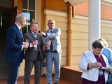 Trzech mężczyzn stoi przed wejściem do szkoły i wznosi toast kieliszkiem szampana. Z prawej strony stoją dwie starsze...