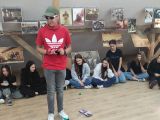 Pośrodku sali stoi chłopak w czerwonej koszulce i czapce z daszkiem na głowie. Na twarzy ma okulary przeciwsłoneczne. Wokól...