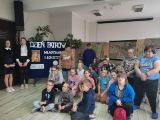 Grupa uczniów siedzi na podłodze w dużej sali. W tle rozwieszone prace plastyczne wykonane na szarym papierze