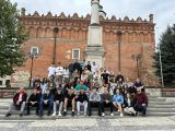 Przed dużym budynkiem z czerwonej cegły stoi duża grupa uczniów liceum wraz z nauczycielami. Między uczniami betonowa...
